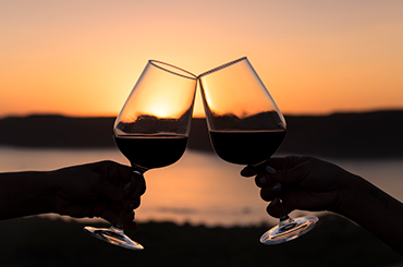 Two hands clinking glasses of red wine at sunset